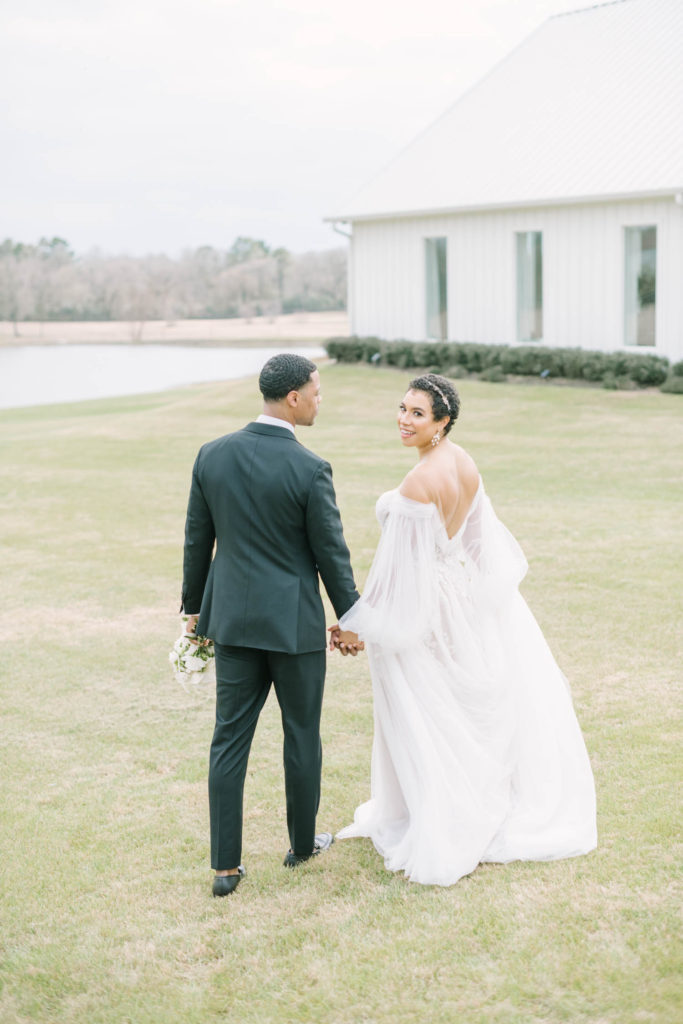 Bride looks back over her shoulder while walking with her groom outside Farmhouse Montgomery in front of a pond by Christina Elliott Photography. over the shoulder pose mr and mrs just married bridal portraits farmhouse weddings Texas #christinaelliottphotography #christinaelliottweddings #TheFarmhouseMontgomery #farmhouseweddings #texasweddings #houstonweddingphotographers #mrandmrs #tildeathdowepart #weddingphotography #tietheknot #Houstonweddings #weddinginspiration #bride #groom