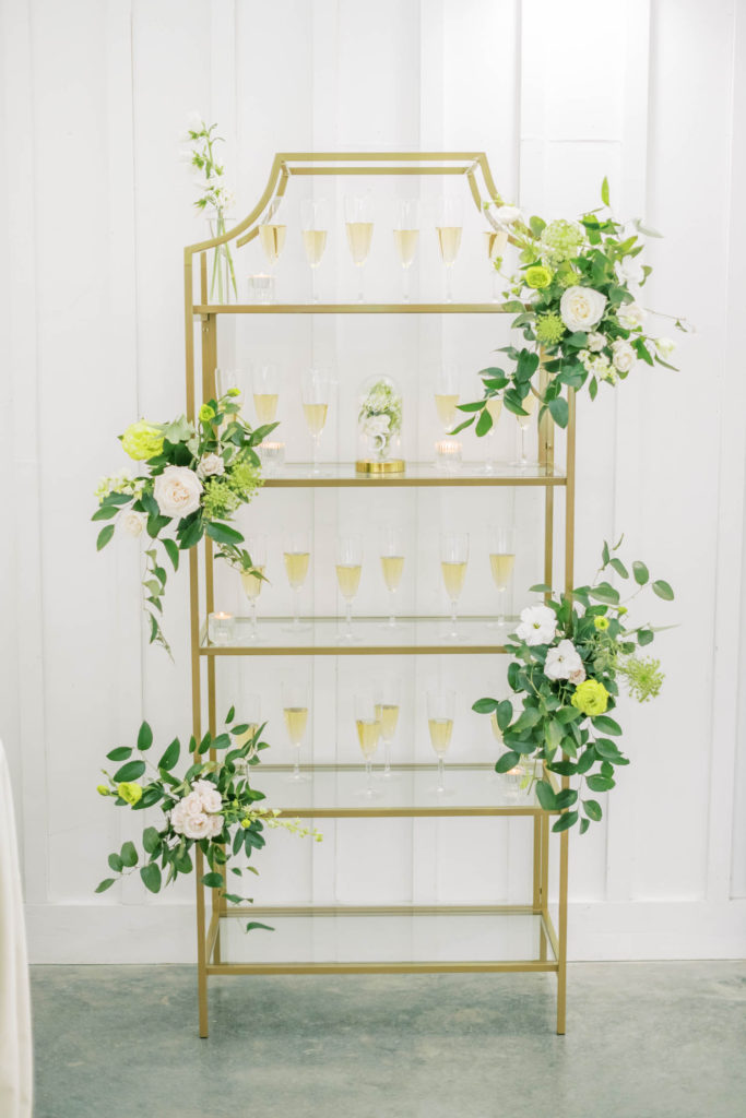On a gold glass bookshelf with floral arrangements, the wedding party has glasses of champagne for toasting displayed by Christina Elliott Photography, a Houston wedding photographer. champagne wedding toast display #christinaelliottphotography #christinaelliottweddings #TheFarmhouseMontgomery #farmhouseweddings #texasweddings #houstonweddingphotographers #mrandmrs #tildeathdowepart #weddingphotography #tietheknot #Houstonweddings #weddinginspiration #bride #groom