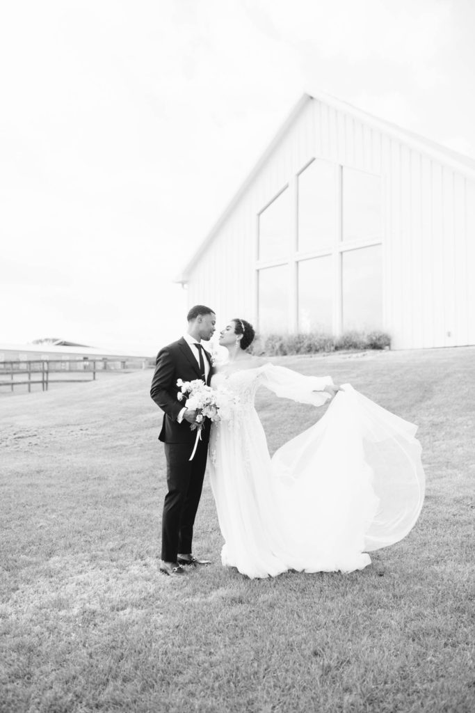 A black and white photo of newly married couple outside of big white farmhouse with bride holding her wedding dress out by Christina Elliott Photography in Houston Texas area. black and white wedding photography bride holding wedding train #christinaelliottphotography #christinaelliottweddings #TheFarmhouseMontgomery #farmhouseweddings #texasweddings #houstonweddingphotographers #mrandmrs #tildeathdowepart #weddingphotography #tietheknot #Houstonweddings #weddinginspiration #bride #groom
