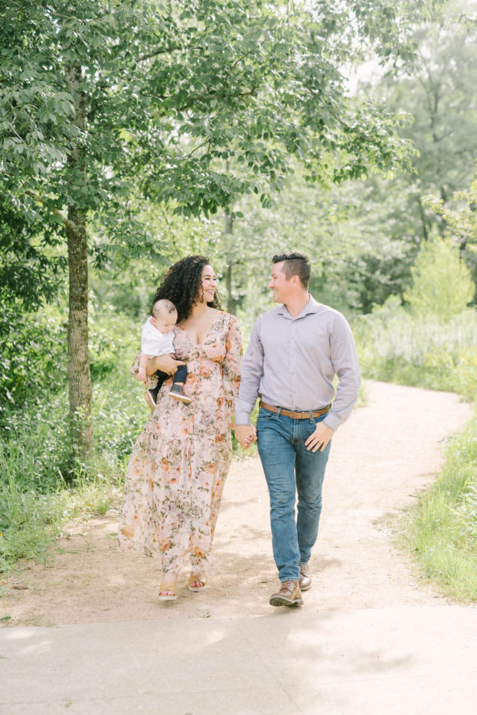 Christina Elliott Photography captures a family of three holding hands, looking at each other and walking on a dirt path at the Houston Arboretum. famioy of three young family pictures family pictures with baby summer family pictures #christinaelliottphotography #christinaelliottfamilies #houstonfamilyphotographer #familyofthree #Houstonarboretum #babyboy #familypictures #outdoorfamilypictures #summerfamilypics #familyphotos #famiyphotographer #Houstonfamilyphotos
