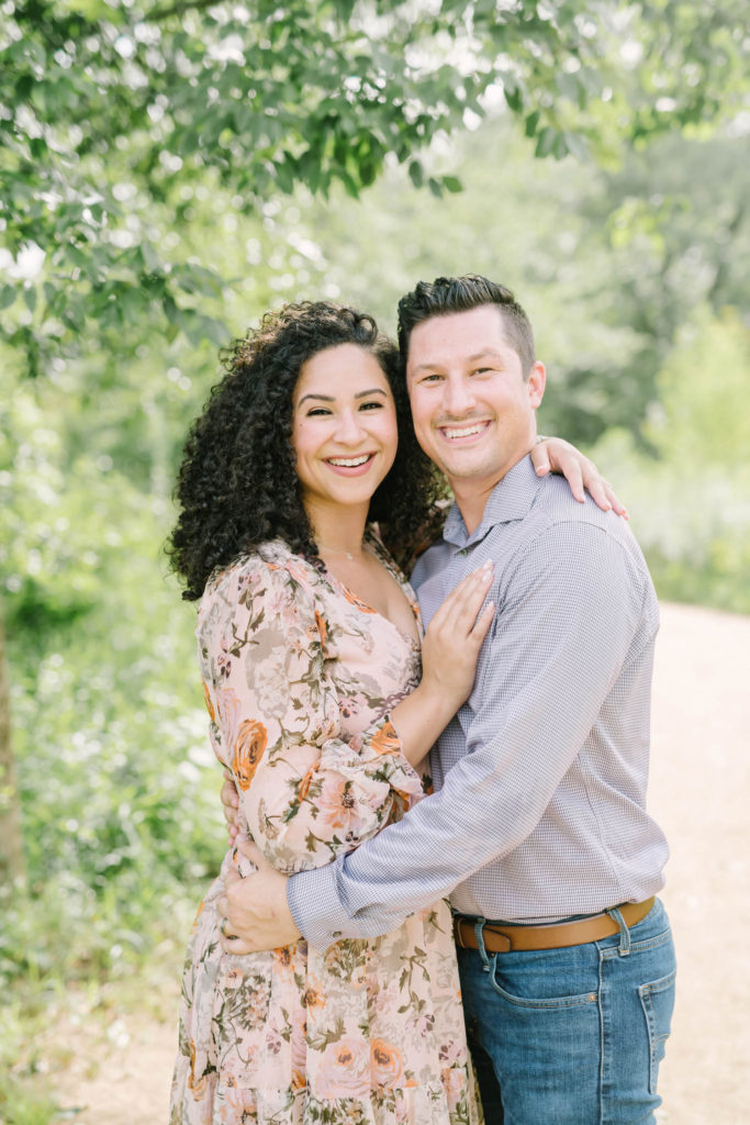 Under a green tree in the summertime a husband and wife smile with their arms wrapped around one another at the Houston Arboretum by Christina Elliott Photography. light pink and orange dress summer family outfit ideas #christinaelliottphotography #christinaelliottfamilies #houstonfamilyphotographer #familyofthree #Houstonarboretum #babyboy #familypictures #outdoorfamilypictures #summerfamilypics #familyphotos #famiyphotographer #Houstonfamilyphotos