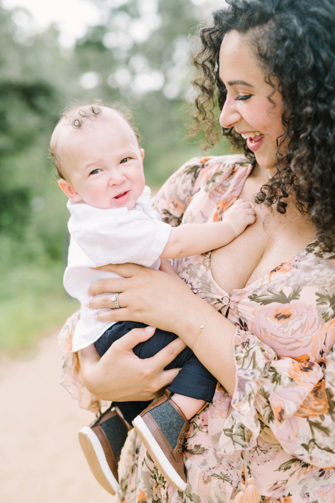 Houston Family Photographer Christina Elliott Photography captures a mother in a light pink dress smiling at her baby boy as she holds him on her arm. houston family photographers baby boy with curly hair baby's first family pic #christinaelliottphotography #christinaelliottfamilies #houstonfamilyphotographer #familyofthree #Houstonarboretum #babyboy #familypictures #outdoorfamilypictures #summerfamilypics #familyphotos #famiyphotographer #Houstonfamilyphotos
