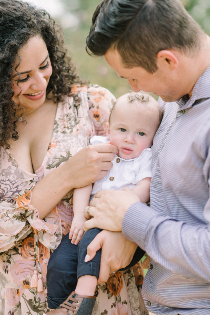At the center of a picture a baby boy with curly brown hair gets tended to by his mother and father in Houston by Christina Elliott Photography. center of the world parents love tender parent moment #christinaelliottphotography #christinaelliottfamilies #houstonfamilyphotographer #familyofthree #Houstonarboretum #babyboy #familypictures #outdoorfamilypictures #summerfamilypics #familyphotos #famiyphotographer #Houstonfamilyphotos