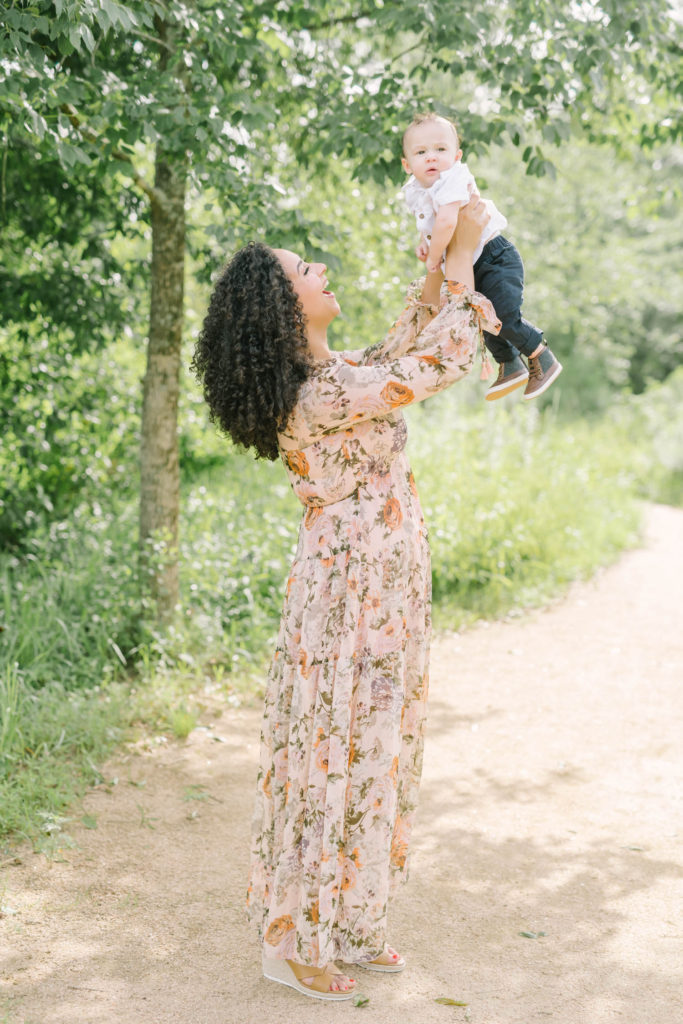 Mother in a light orange dress holds her baby boy up in the air and smiles at him by Christina Elliott Photography in Houston, Texas. baby boy in brown boots white button down baby shirt mother and baby boy #christinaelliottphotography #christinaelliottfamilies #houstonfamilyphotographer #familyofthree #Houstonarboretum #babyboy #familypictures #outdoorfamilypictures #summerfamilypics #familyphotos #famiyphotographer #Houstonfamilyphotos