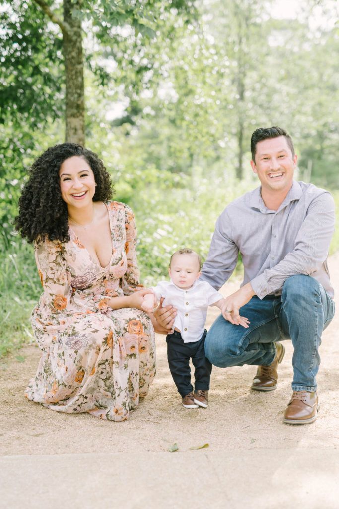 Family Photographer captures a woman with beautiful curly hair and a stylish husband crouched down holding their babies hands in Houston by Christina Elliott Photography. family of three photogenic family photos Houston Photography #christinaelliottphotography #christinaelliottfamilies #houstonfamilyphotographer #familyofthree #Houstonarboretum #babyboy #familypictures #outdoorfamilypictures #summerfamilypics #familyphotos #famiyphotographer #Houstonfamilyphotos