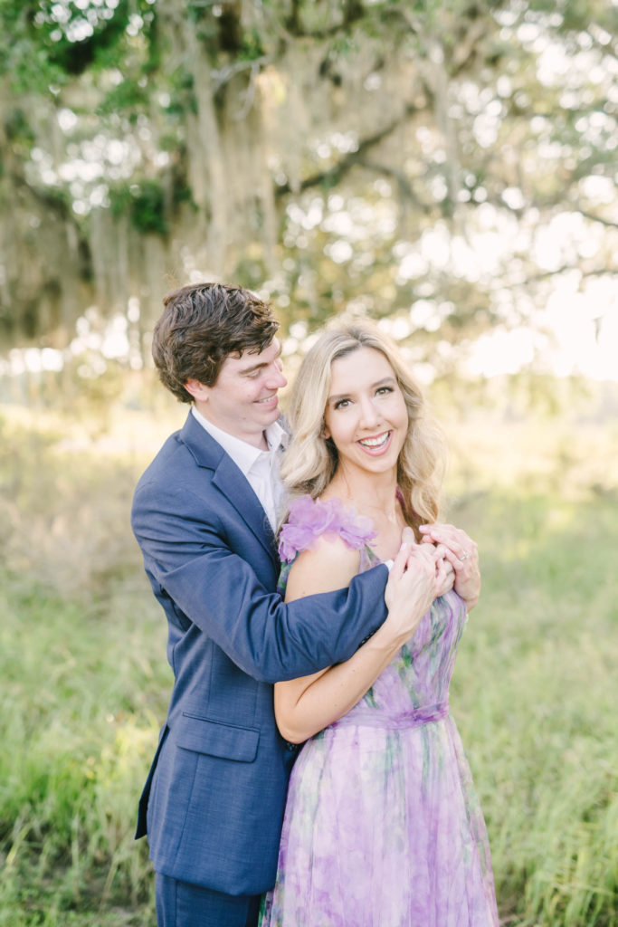 Bride-to-be smiles as her fiance wraps his arms around her from behind and smiles down at her by Christina Elliott Photography in Houston, Texas. unique engagement style green grass field mossy trees #christinaelliottphotography #christinaelliottengagements #houstonengagements #brazosbendstateparkengagements #brazosbendphotography #houstonengagementphotographer #weddingannoucementphotos #houstonphotographers #couplegoals #soontobemarried #engaged #engagementphotos