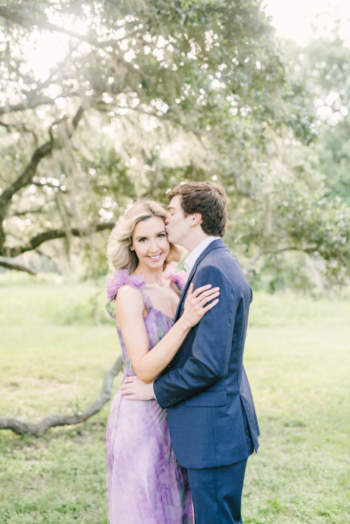A man kisses his soon-to-be wife's head as she snuggles close at Brazos Bend State Park in Houston with Christina Elliott Photography. purple engagement dress kissing couple poses #christinaelliottphotography #christinaelliottengagements #houstonengagements #brazosbendstateparkengagements #brazosbendphotography #houstonengagementphotographer #weddingannoucementphotos #houstonphotographers #couplegoals #soontobemarried #engaged #engagementphotos