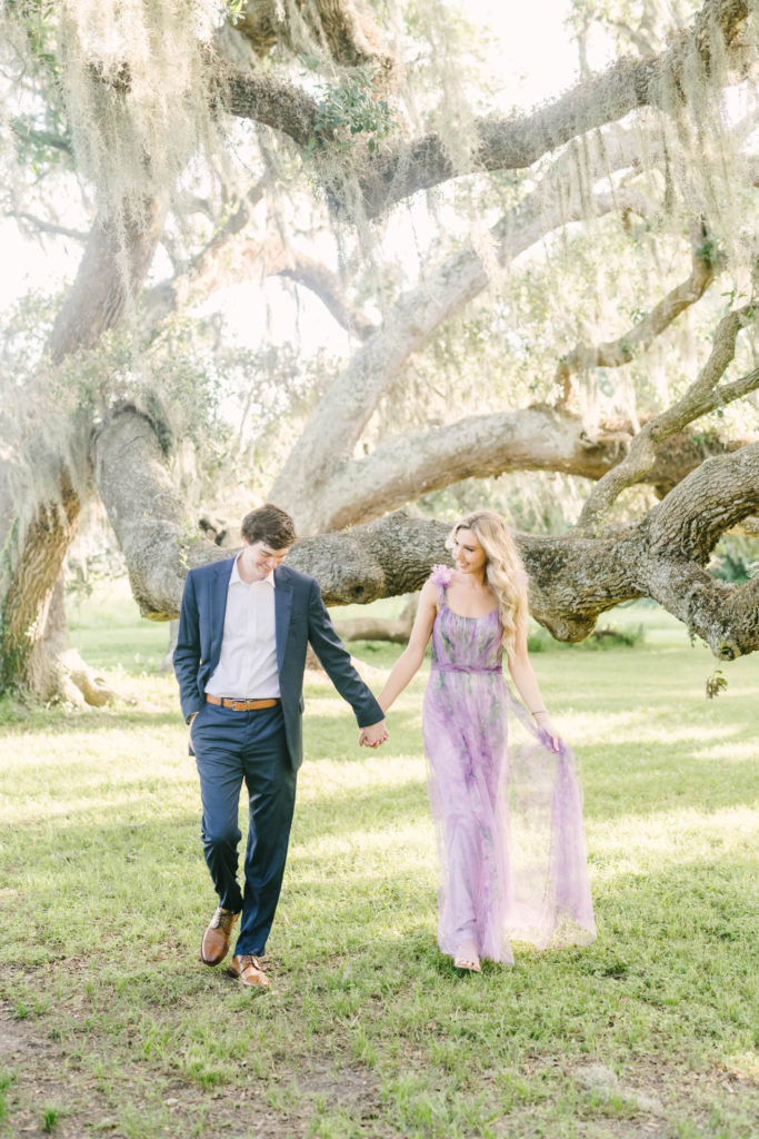 At Brazos Bend State Park a couple walks hand in hand in front of a mossy oak tree with a woman wearing a purple chiffon gown by Christina Elliott Photography. walking hand in hand pictures Houston engagement photographers #christinaelliottphotography #christinaelliottengagements #houstonengagements #brazosbendstateparkengagements #brazosbendphotography #houstonengagementphotographer #weddingannoucementphotos #houstonphotographers #couplegoals #soontobemarried #engaged #engagementphotos