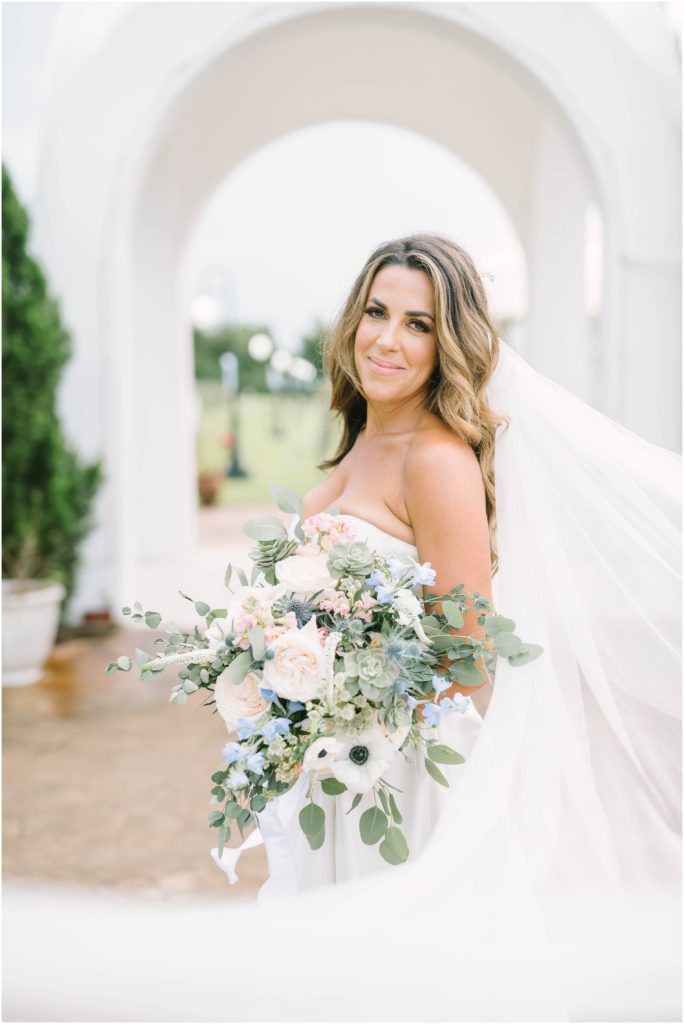 Beautiful bride stands underneath a white arch and smiles in a strapless wedding gown with a stunning muted floral bouquet and veil spread out by Christina Elliott Photography in Houston. bridal portrait bridal veil portraits soft colors #christinaelliottphotography #christinaelliottweddings #Winerywedding #Houstonweddings #newlyweds #weddingdress #weddingphotography #Houstonweddingphotographers #dreamyweddings #bluesuit #weddingstyle #mrandmrs #weddinggoals #Houstonphotographers