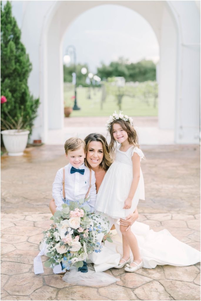 At a Houston Winery aa bride sits down and smiles with her two kids her little girl in a white dress and little boy in a blue bow tie by Christina Elliott Photography. bride and her kids mommy bridal picture kids in weddings #christinaelliottphotography #christinaelliottweddings #Winerywedding #Houstonweddings #newlyweds #weddingdress #weddingphotography #Houstonweddingphotographers #dreamyweddings #bluesuit #weddingstyle #mrandmrs #weddinggoals #Houstonphotographers