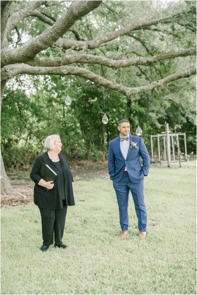 A groom waits with wedding officiant under a big tree with glass candle globes at the end of the aisle for his bride captured by Christina Elliott Photography a Houston photographer. glass candle globes for wedding groom at end of aisle #christinaelliottphotography #christinaelliottweddings #Winerywedding #Houstonweddings #newlyweds #weddingdress #weddingphotography #Houstonweddingphotographers #dreamyweddings #bluesuit #weddingstyle #mrandmrs #weddinggoals #Houstonphotographers