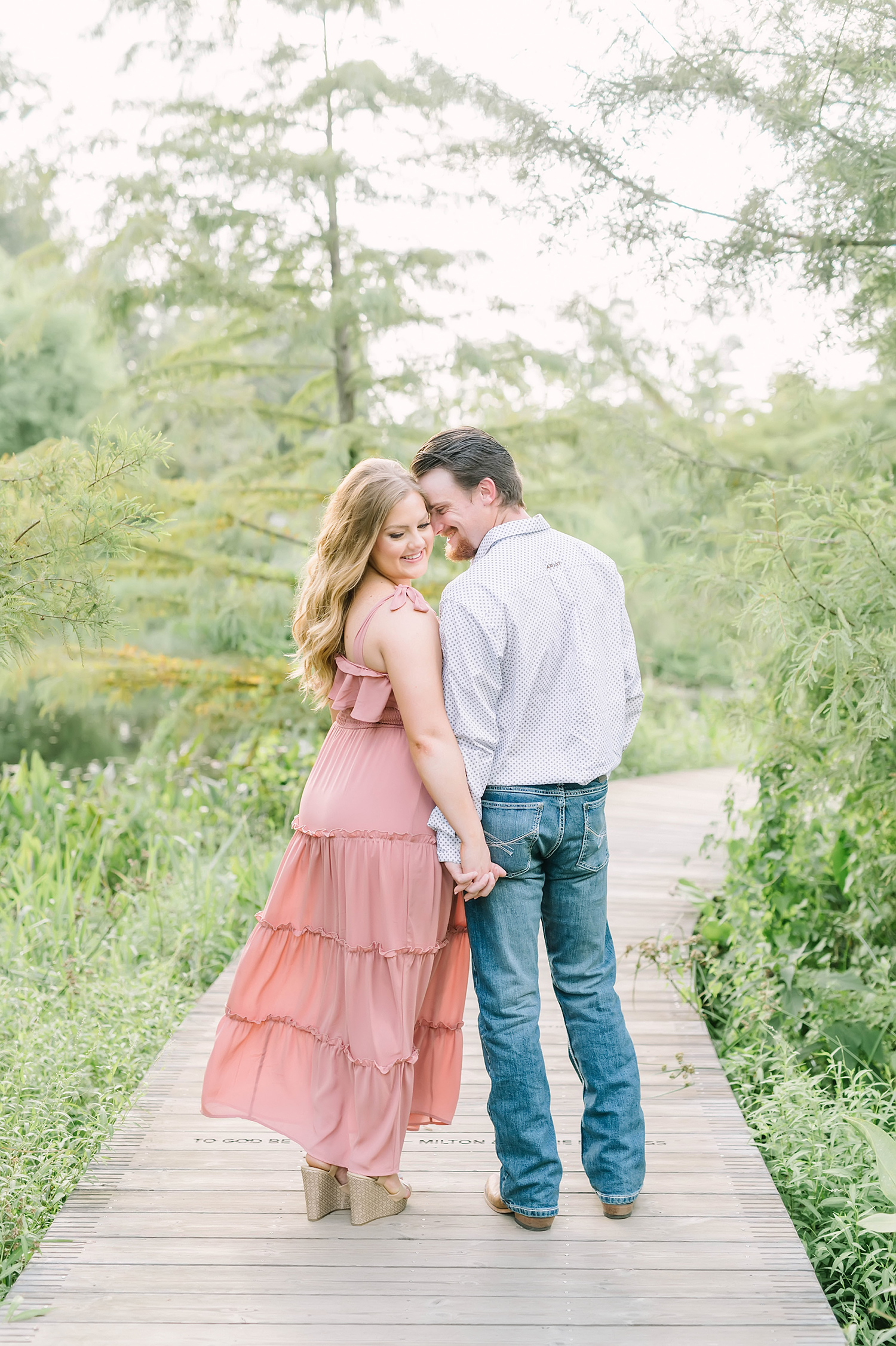 A woman in a pink dress holds hands and stands on a bridge with her fiance as she looks over her shoulder in Houston, Texas by Christina Elliott Photography. cowboy engagement pictures pink bow tie dress for pic engagement pose ideas wedding annoucement photos #christinaelliottphotography #christinaelliottengagement #houstonengagement #houstonphotographers #houstonarboretum #stillwaterranchwedding #engagementphotos #soontobemarried #outdoorengagments #sayIdo #southernengagements