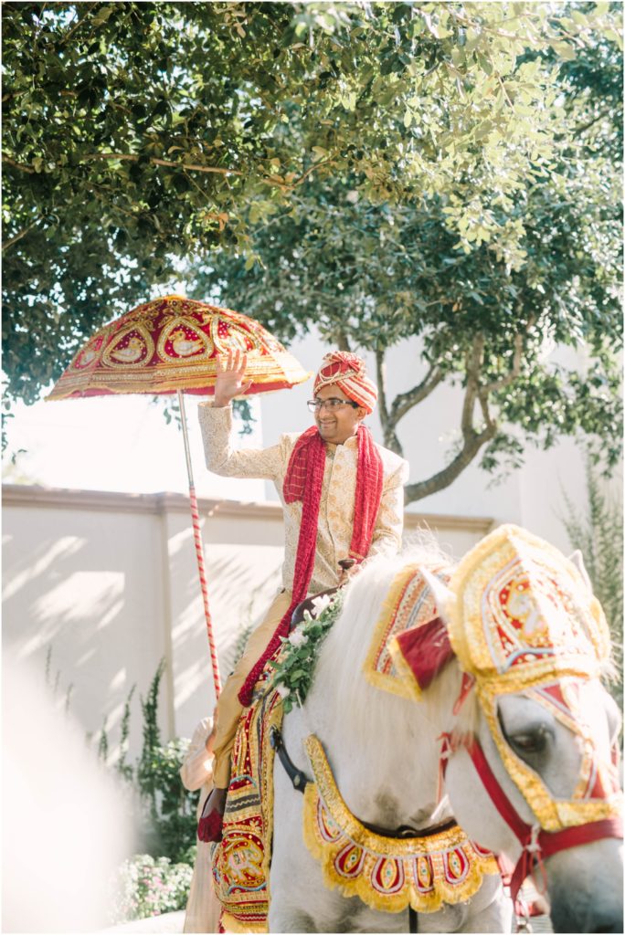 An Indian groom wearing traditional wedding attire rides in on a horse decorated in gold and red with a detailed umbrella by Christina Elliott Photography a Houston photographer. traditional groom on horse for Indian culture #christinaelliottphotography #christinaelliottweddings #thebelltoweron34th #belltowerwedding #houstonweddings #houstonweddingphotographers #mrandmrs #newlymarried #weddinginspiration #bridalgown #groomstyle #texasweddingphotographers #tietheknot #traditionalindianwedding
