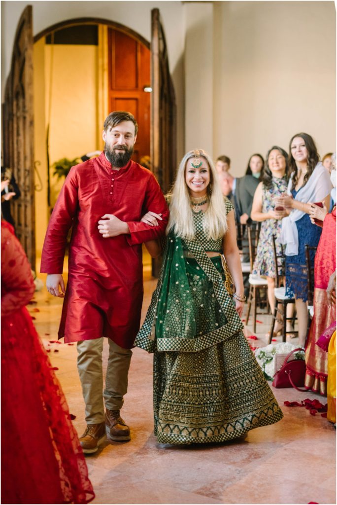 A blonde woman in a traditional emerald green Indian wedding gown is escorted down the aisle with a big smile on her face by Houston photographer Christina Elliott Photography. Emerald green and gold wedding dress #christinaelliottphotography #christinaelliottweddings #thebelltoweron34th #belltowerwedding #houstonweddings #houstonweddingphotographers #mrandmrs #newlymarried #weddinginspiration #bridalgown #groomstyle #texasweddingphotographers #tietheknot #traditionalindianwedding