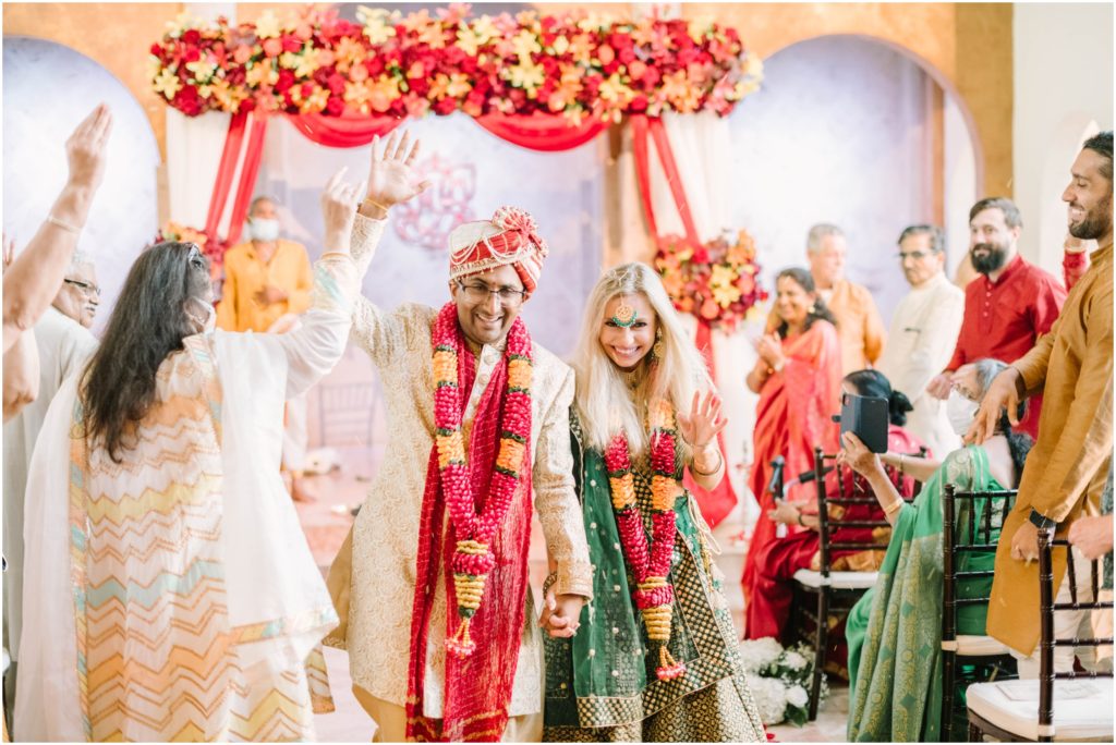 At a colorful wedding, Christina Elliott Photography captures the bride and groom holding hands and leaving the Indian Chapel in Houston, Texas. Bride and Groom in Traditional Indian wedding attire #christinaelliottphotography #christinaelliottweddings #thebelltoweron34th #belltowerwedding #houstonweddings #houstonweddingphotographers #mrandmrs #newlymarried #weddinginspiration #bridalgown #groomstyle #texasweddingphotographers #tietheknot #traditionalindianwedding