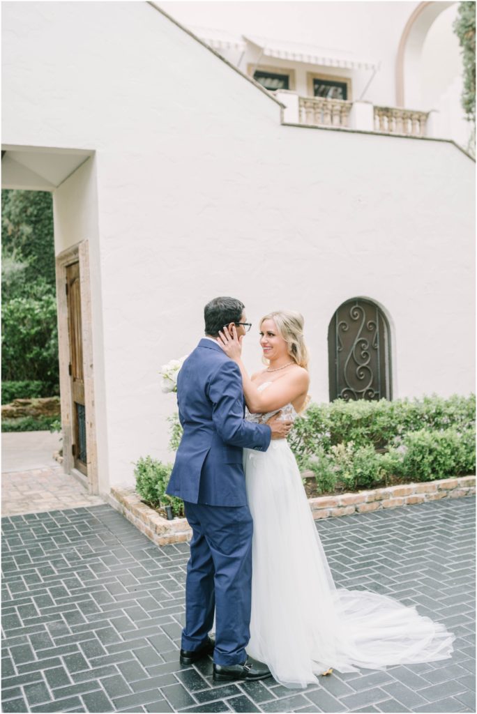 Bride holds the groom's face as he holds her close as he gets the first look at her in a white wedding gown by Christina Elliott Photography in Houston, Texas. bride in love first look photo ideas #christinaelliottphotography #christinaelliottweddings #thebelltoweron34th #belltowerwedding #houstonweddings #houstonweddingphotographers #mrandmrs #newlymarried #weddinginspiration #bridalgown #groomstyle #texasweddingphotographers #tietheknot #traditionalindianwedding