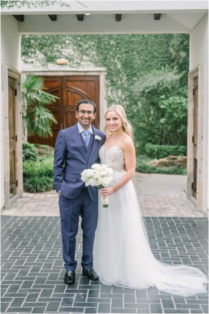 Under a white arch entrance a bride and groom smile in their classy wedding attire in Houston by Christina Elliott Photography. couple wedding portrait beautiful Houston wedding venues #christinaelliottphotography #christinaelliottweddings #thebelltoweron34th #belltowerwedding #houstonweddings #houstonweddingphotographers #mrandmrs #newlymarried #weddinginspiration #bridalgown #groomstyle #texasweddingphotographers #tietheknot #traditionalindianwedding