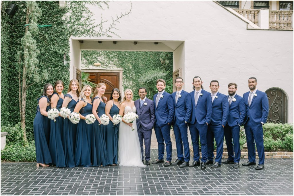 Bride and groom with their wedding party all wearing blue with green vines behind them by Houston photographer Christina Elliott Photography. bridal party wedding colors blue dusty blue wedding colors #christinaelliottphotography #christinaelliottweddings #thebelltoweron34th #belltowerwedding #houstonweddings #houstonweddingphotographers #mrandmrs #newlymarried #weddinginspiration #bridalgown #groomstyle #texasweddingphotographers #tietheknot #traditionalindianwedding