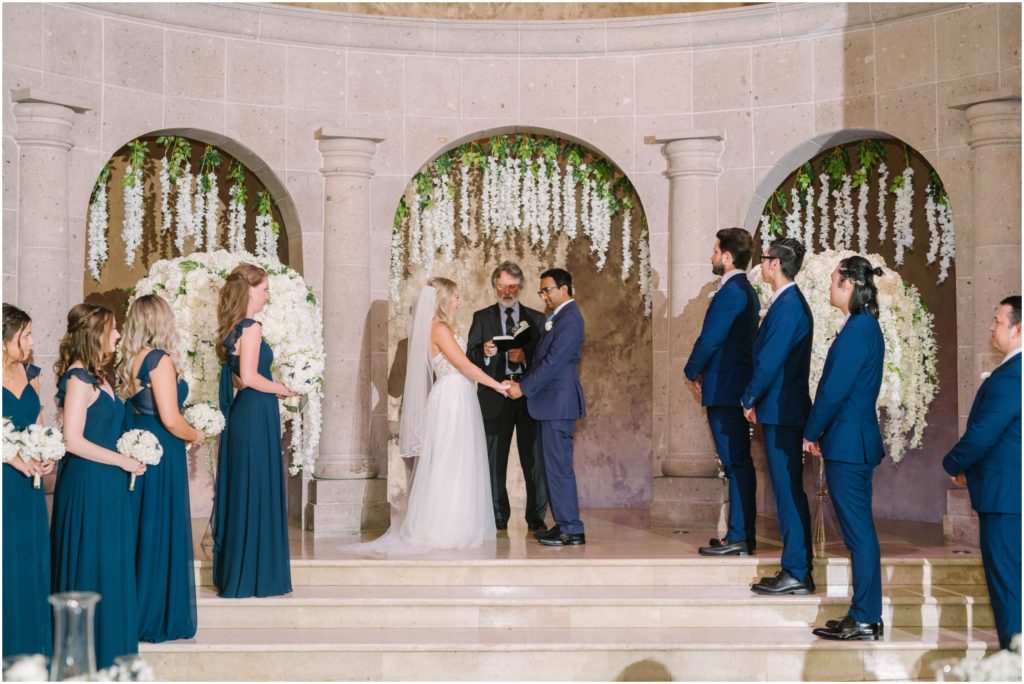 Bride and groom at the end of the aisle with a wedding party on the stairs leading up to them captured by Christina Elliott Photography in Houston, Texas. bride and groom hold hands at altar #christinaelliottphotography #christinaelliottweddings #thebelltoweron34th #belltowerwedding #houstonweddings #houstonweddingphotographers #mrandmrs #newlymarried #weddinginspiration #bridalgown #groomstyle #texasweddingphotographers #tietheknot #traditionalindianwedding