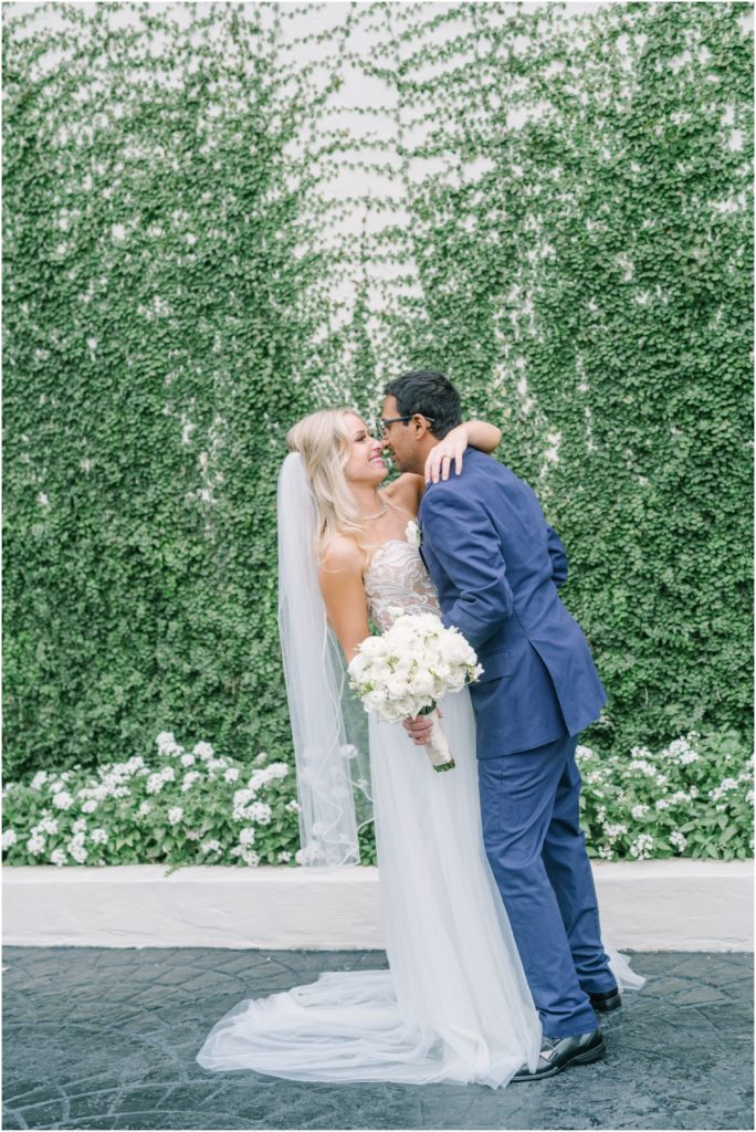 Christina Elliott Photography captures a glowing happy couple going in for a kiss in front of a green vine wall in Houston. married happy couple kissing wedding couple wedding inspiration #christinaelliottphotography #christinaelliottweddings #thebelltoweron34th #belltowerwedding #houstonweddings #houstonweddingphotographers #mrandmrs #newlymarried #weddinginspiration #bridalgown #groomstyle #texasweddingphotographers #tietheknot #traditionalindianwedding