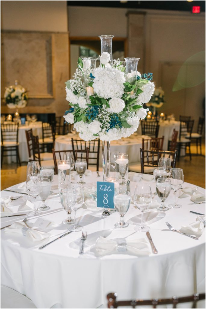 At The Bell Tower on 34th in Houston Christina Elliott Photography captures a round table with tall flower centerpiece in blue and white colors. tall floral centerpiece class flower stand #christinaelliottphotography #christinaelliottweddings #thebelltoweron34th #belltowerwedding #houstonweddings #houstonweddingphotographers #mrandmrs #newlymarried #weddinginspiration #bridalgown #groomstyle #texasweddingphotographers #tietheknot #traditionalindianwedding