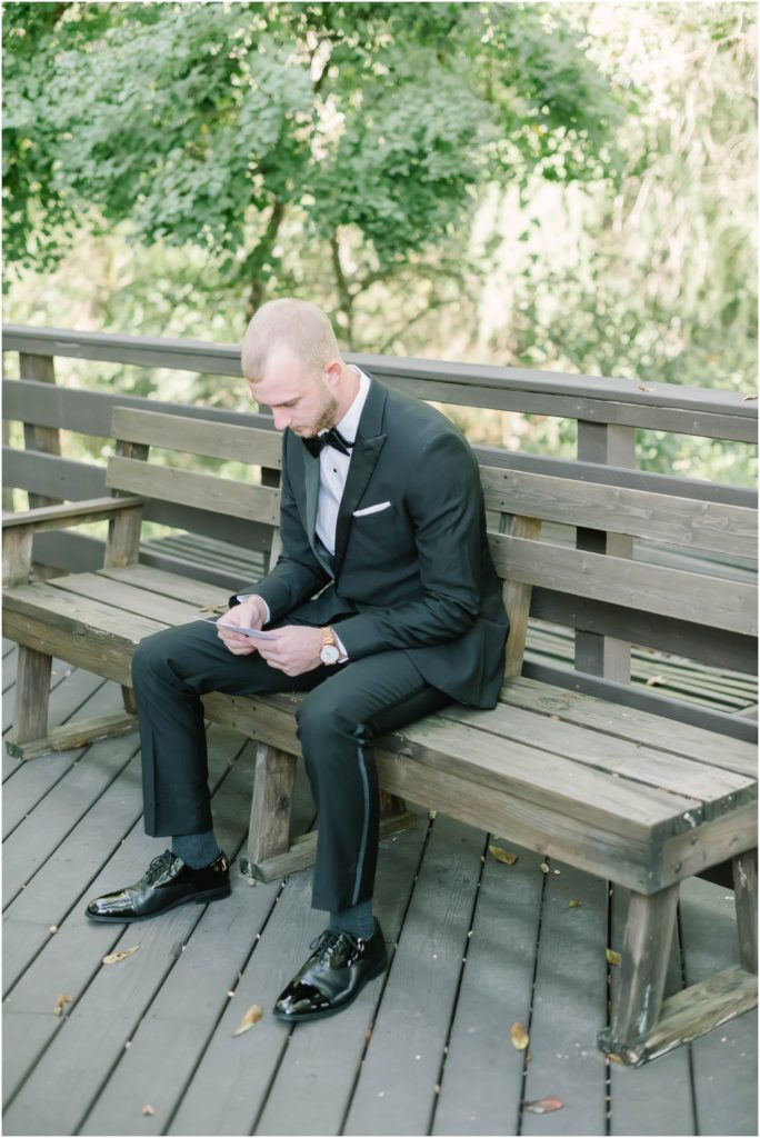 Christina Elliott Photography captures the groom sitting outside on a wooden bench reading a note from the bride before the wedding in Houston, Texas. groom reading note from bride wedding detail ideas #christinaelliottphotography #christinaelliottweddings #houstontx #houstonweddings #houstonweddingphotographers #carriagehousewedding #conroetexas #texasweddingvenues #carriagehousechapel #cottageweddings #farmhousewedding #sweatheartweddinggown #countrywedding #weddinginspiration