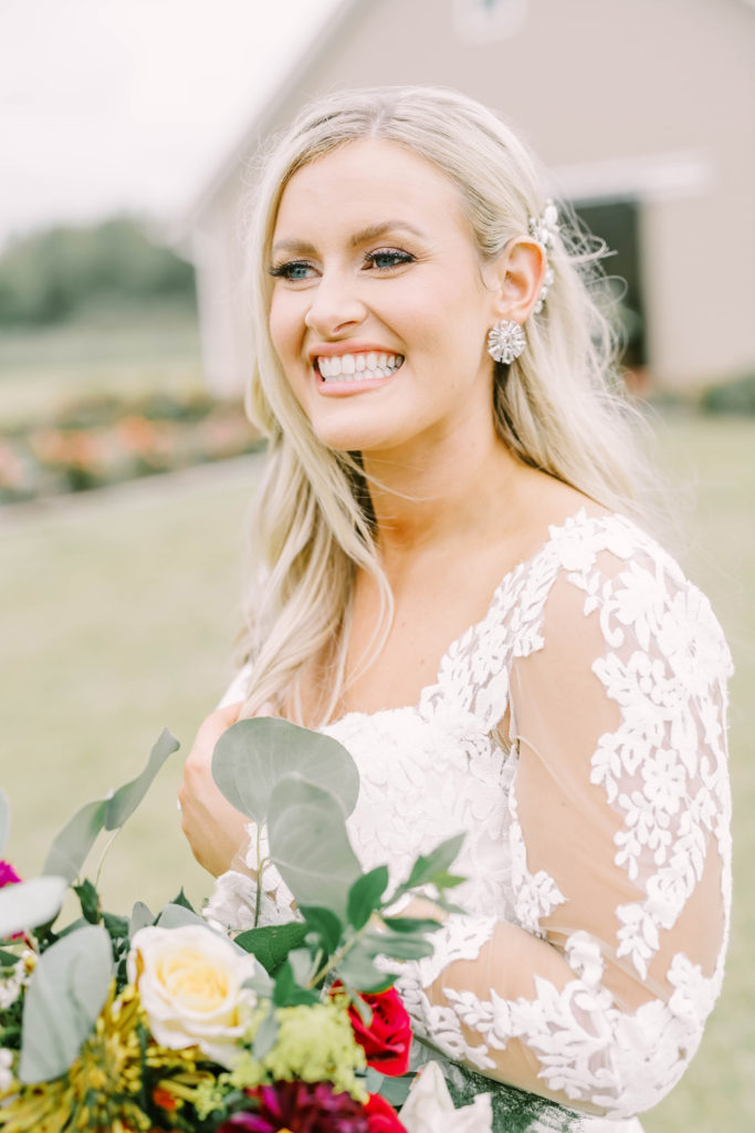 A Texas wedding photographer captures a bride with a big smile wearing a sheer lace sleeves wedding dress. bridal details add on sleeves to wedding dress #christinaelliottphotography #texasweddingphotographer #ranchwedding #countrychic