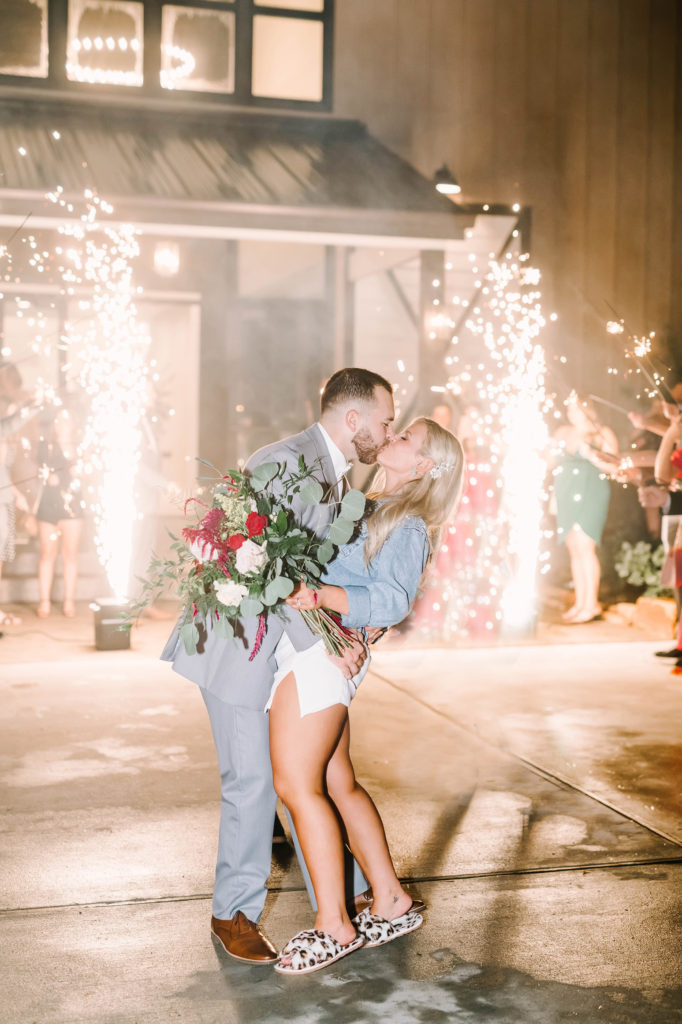 With sparklers behind the newlyweds, they kiss as they dress more casually and get ready to leave by Christina Elliott Photography. wedding send off send off attire #christinaelliottphotography #texasweddingphotographer #ranchwedding #countrychic