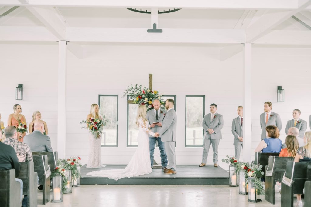 At a Texas wedding venue, a couple gets married with a floral cross altar and vaulted ceilings by Christina Elliott Photography. bridal party upfront guests in pews #christinaelliottphotography #texasweddingphotographer #ranchwedding #countrychic