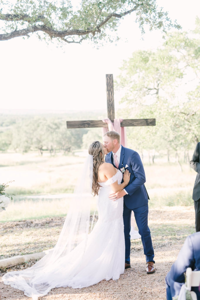 Bride and groom kiss at the alter decorated with a rustic wooden cross and a mauve drapery