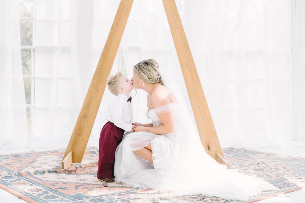 Bride kisses her toddler son during her wedding by Christina Elliott Photography in Texas. toddler kisses his mother bride off the should dress #christinaelliottphotography #theoakatelierwedding #Houstonwinterweddings #weddingphotographersHouston