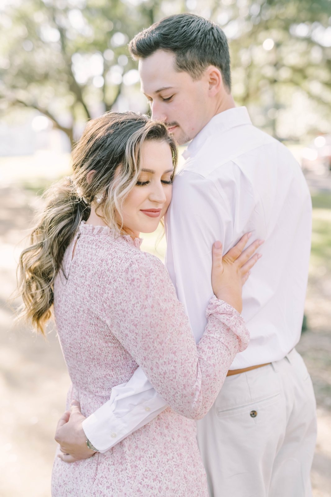 Wearing a pink floral dress in the summer sunlight a woman holds onto her fiance by Christina Elliott Photography. pink dress #christinaelliottphotography #Houstonengagements #riceuniversity #springengagements #sayIdo #Houstonengagementphotographers