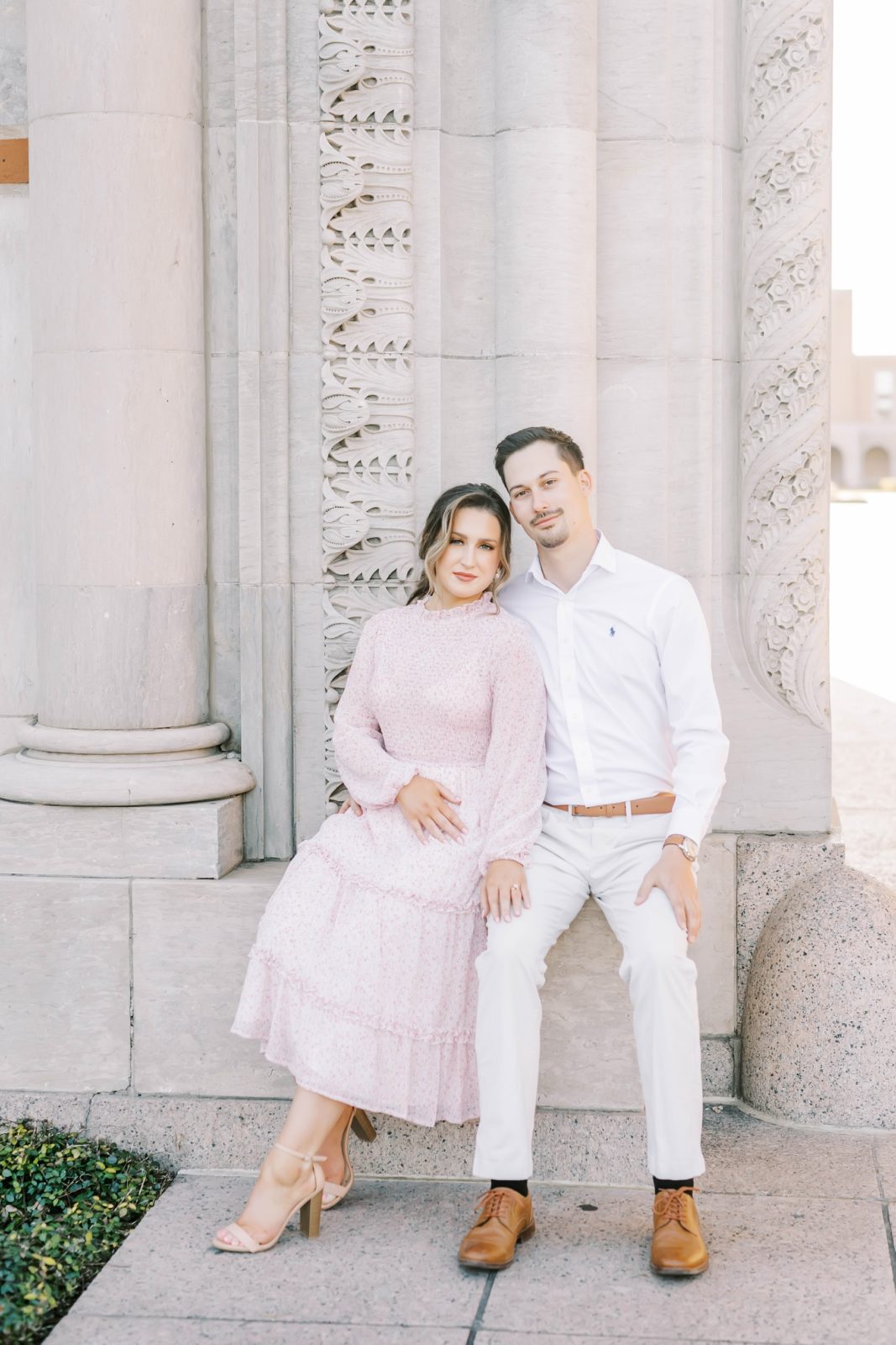 Man in all white holds his fiance in a pink gown close while sitting by Christina Elliott Photography. brown shoes ponytail #christinaelliottphotography #Houstonengagements #riceuniversity #springengagements #sayIdo #Houstonengagementphotographers