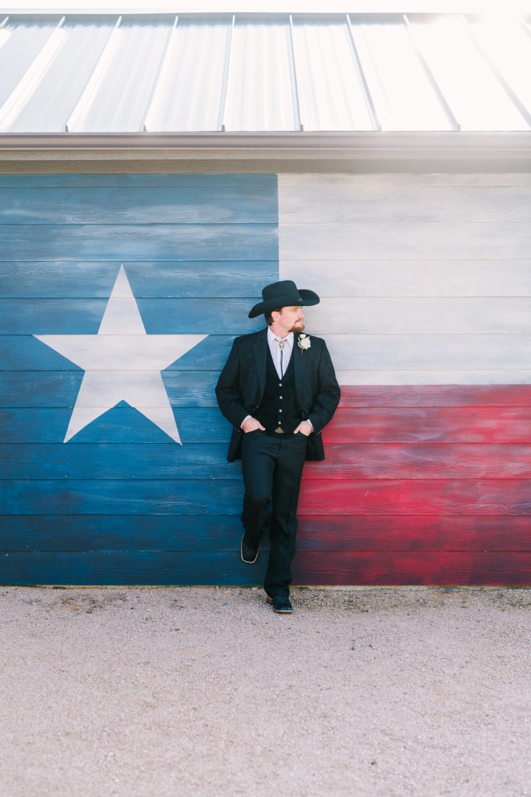 Groom wearing black cowboy attire for a wedding is in front of the Texas flag by Christina Elliott Photography. texas groom #ChristinaElliottPhotography #ChristinaElliottWeddings #StillWatersRanchWedding #Texasweddings #countrywedding #ranchwedding