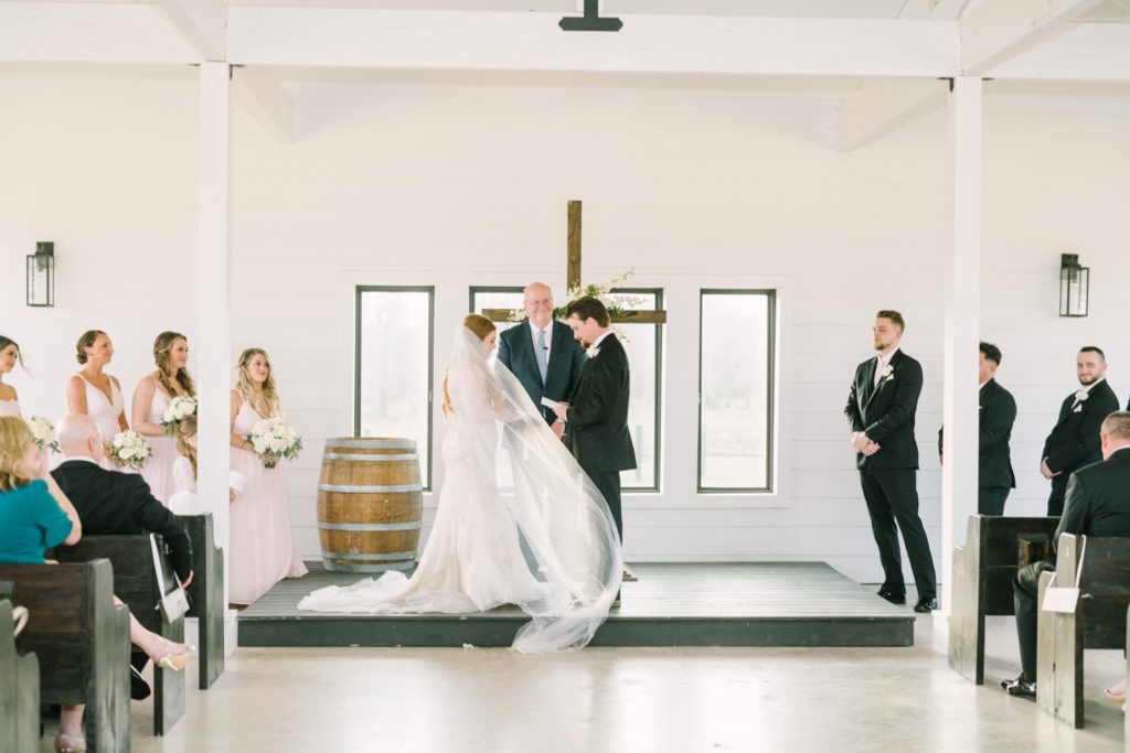 Bride and groom stand at an altar with wooden cross by Christina Elliott Photography. wooden cross altar decor veil in wind #ChristinaElliottPhotography #ChristinaElliottWeddings #StillWatersRanchWedding #Texasweddings #countrywedding #ranchwedding