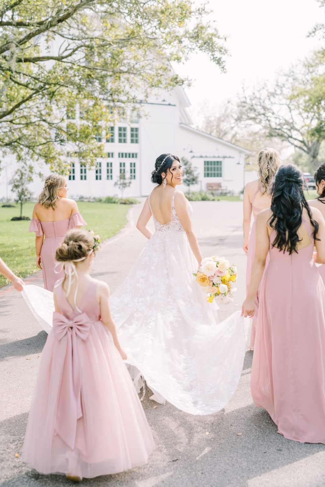Surrounded by bridesmaids a bride looks back over her shoulder while walking toward a white barn by Christina Elliott Photography. lace dress #ChristinaElliottPhotography #ChristinaElliottWeddings #Houstonwedding #TheSpringsVenue #EastHoustonwedding