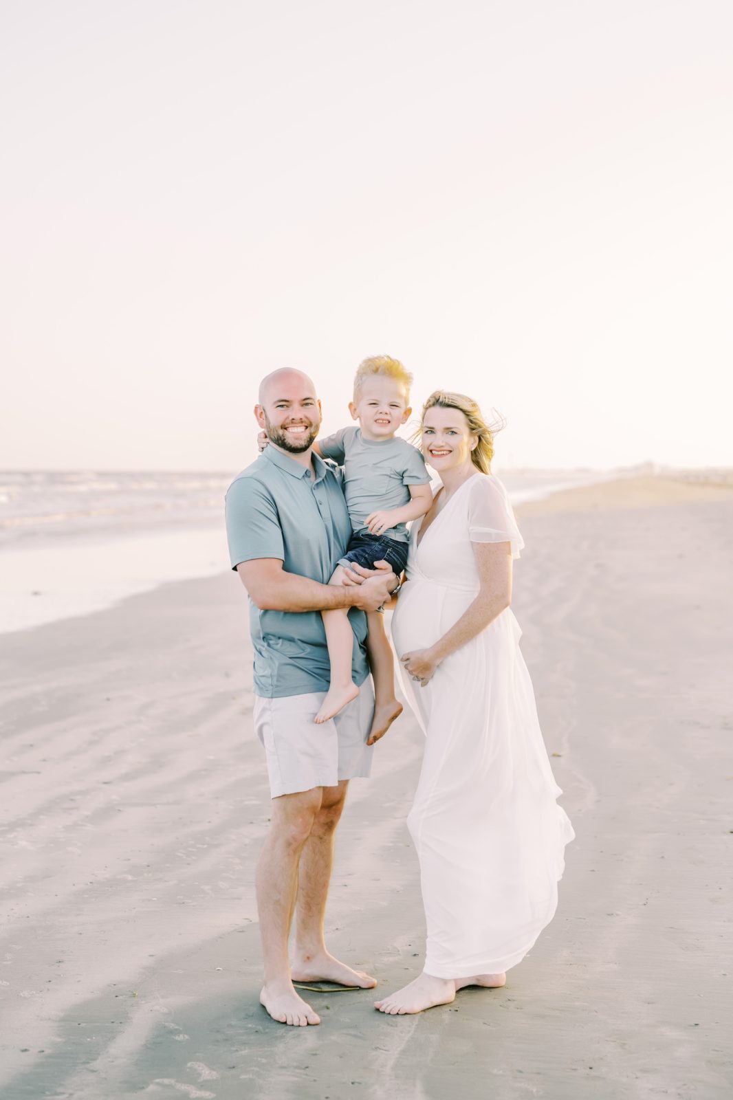 Standing on the beach in Galveston a family smiles together captured by Christina Elliott Photography. boy baby on way #ChristinaElliottPhotography #ChristinaElliottFamilies #GalvestonPhotography #Beachmaternityphotography #Texasfamilyphotographers