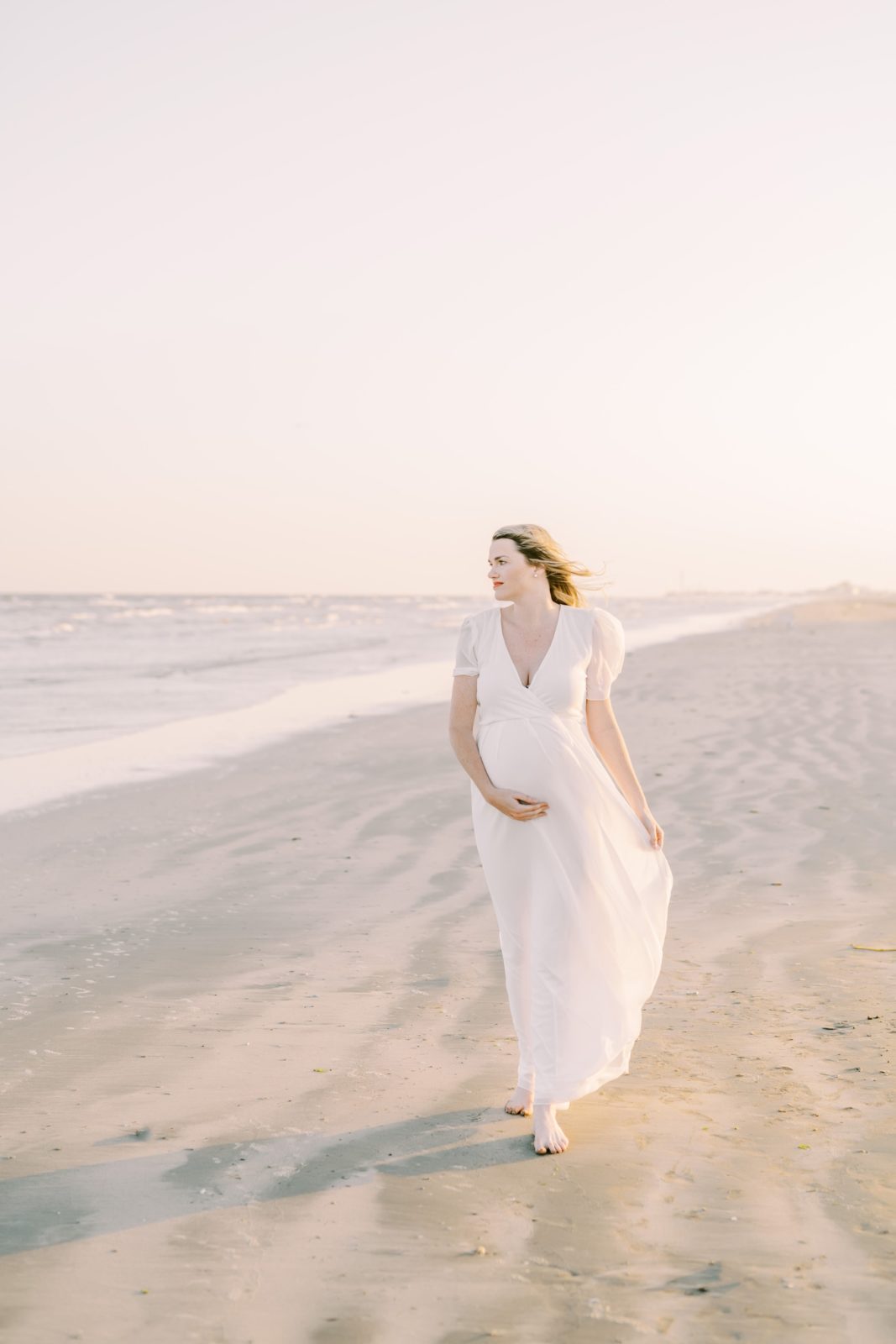 Timeless maternity portrait of woman walking along the beach by Christina Elliott Photography. white maternity dress #ChristinaElliottPhotography #ChristinaElliottFamilies #GalvestonPhotography #Beachmaternityphotography #Texasfamilyphotographers