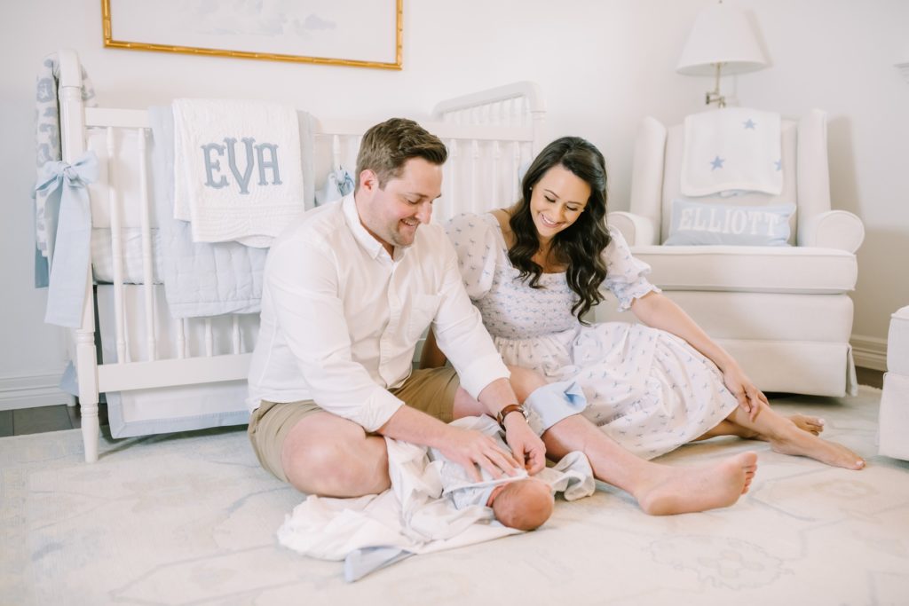 Mother and father sit on the floor of a nursery and swaddle their baby girl by Christina Elliott Photography. blue nursery #ChristinaElliottPhotography #ChristinaElliottNewborns #HoustonLifestyleNewborns #HoustonFamilyPhotographer