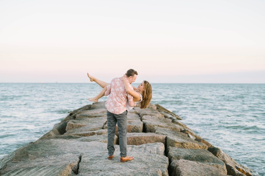 On a rocky jetty in the ocean a man holds his fiance in his arm and dips her for a kiss by Christina Elliott Photography. Ocean engagements #ChristinaElliottPhotography #ChristinaElliottEngagements #TheTremontHouse #Texasengagements #shesaidyes