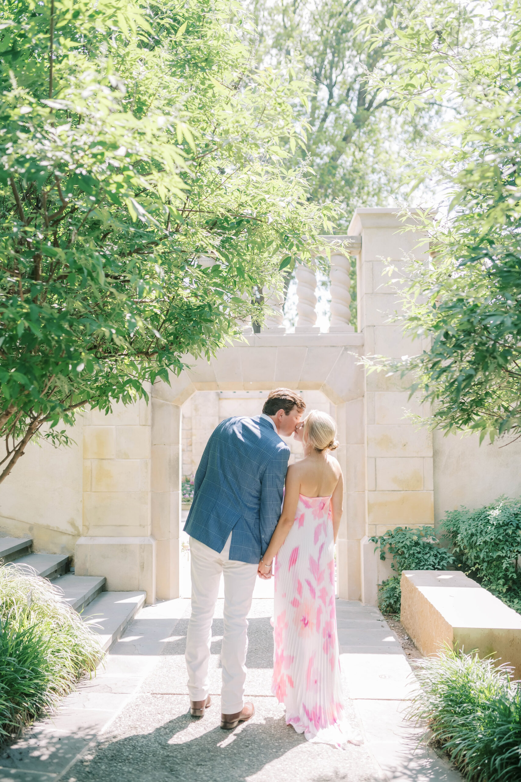 A couple stands together in a garden, the man in a blue jacket and white pants, and the woman in a pink and white dress. They are both smiling and surrounded by greenery and flowers at the dallas arboretum
